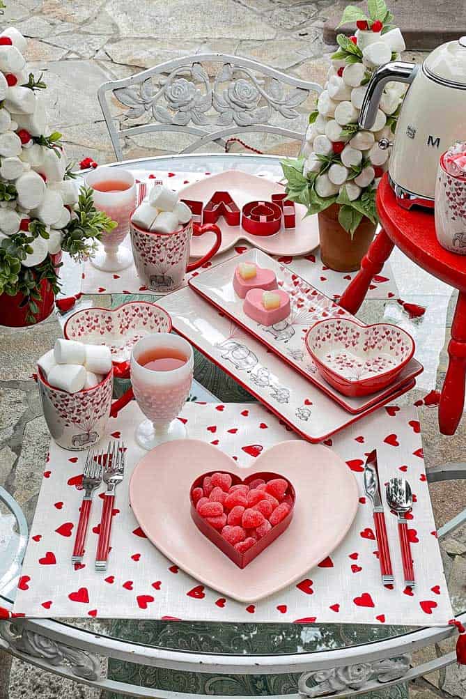 A romantic table setting features heart-shaped plates, red and white dishware, drinks, and marshmallow treats on a white tablecloth with red hearts—perfect inspiration for Valentine’s Day Dinner Table Decoration Ideas, complete with flowers and sweet accents.