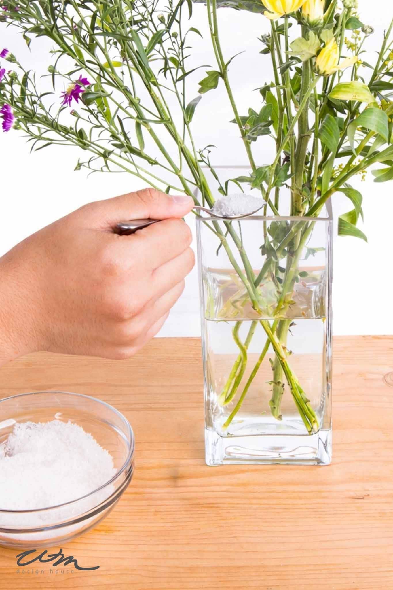 A hand adds white powder from a spoon into a glass vase of fresh flowers with water—showing how to keep flower arrangements fresh. A bowl of the same powder sits on the wooden table nearby.