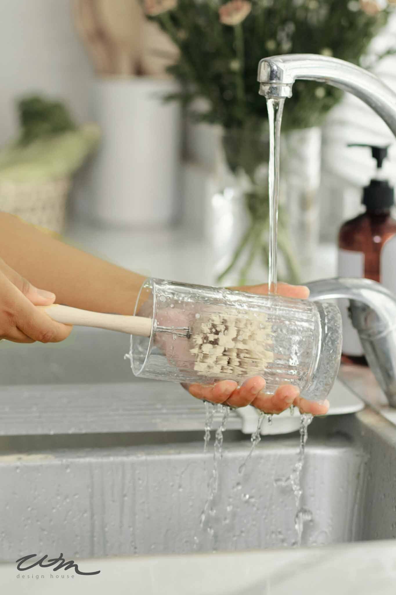 How to Keep Flower Arrangements Fresh and Beautiful for Days. A person washes a clear glass with a brush under running water at a kitchen sink, water splashing over their hand. Blurred flowers and a soap dispenser appear in the background, evoking tips on how to keep flower arrangements fresh. 