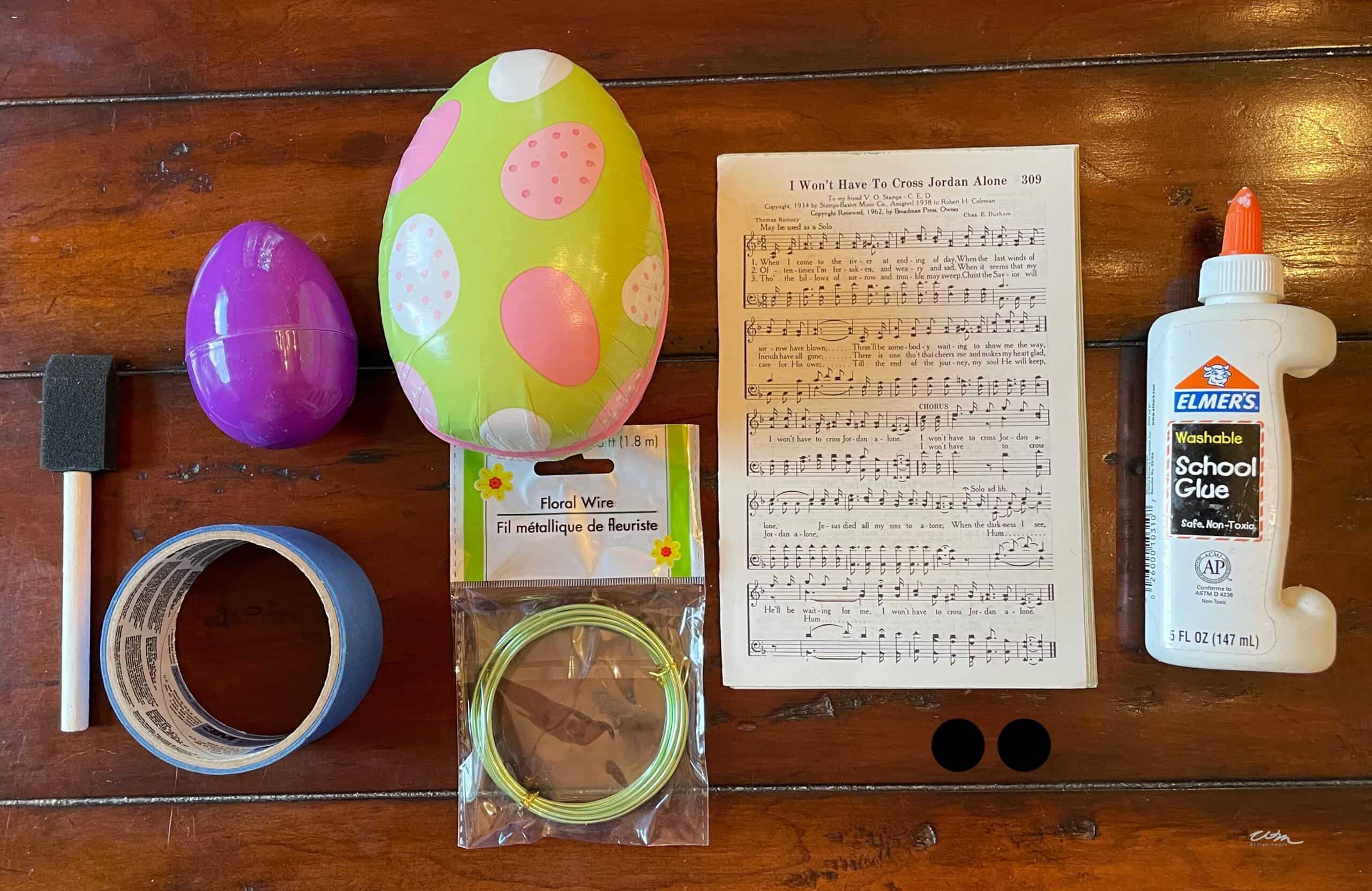 Art supplies for a Paper Mache Easter Bunny on a wooden table: two plastic Easter eggs, foam brush, blue painter’s tape, floral wire, sheet music, two black circles, and Elmer’s washable school glue.