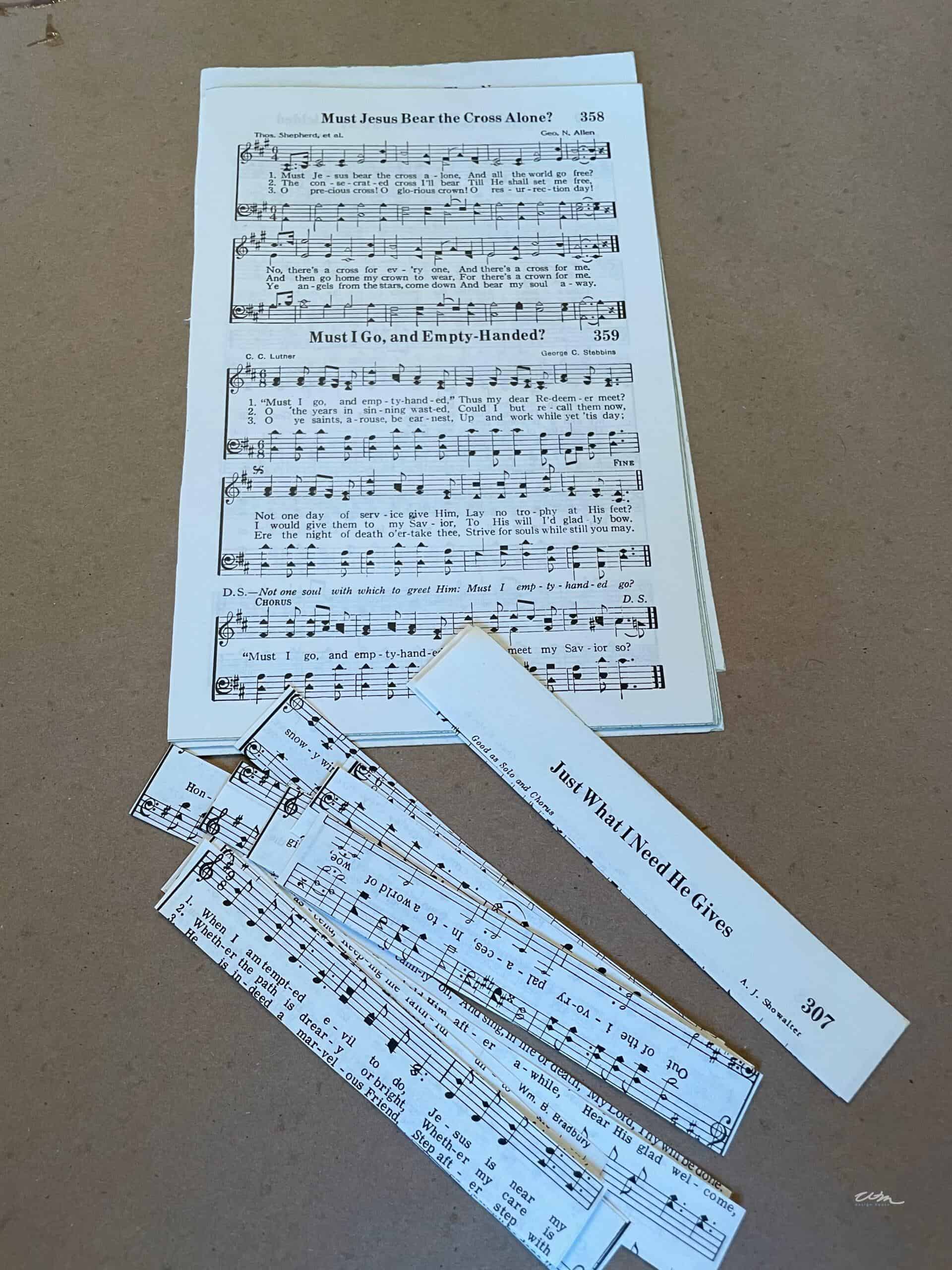 A hymnal page with sheet music and lyrics sits on a brown surface, along with several paper strips cut from similar hymnal pages, and a rectangular piece labeled "Just What I Need He Gives.