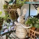 A decorative bunny figurine wrapped in sheet music paper with a ribbon around its neck sits on a shelf surrounded by potted plants, greenery, and garden decor.