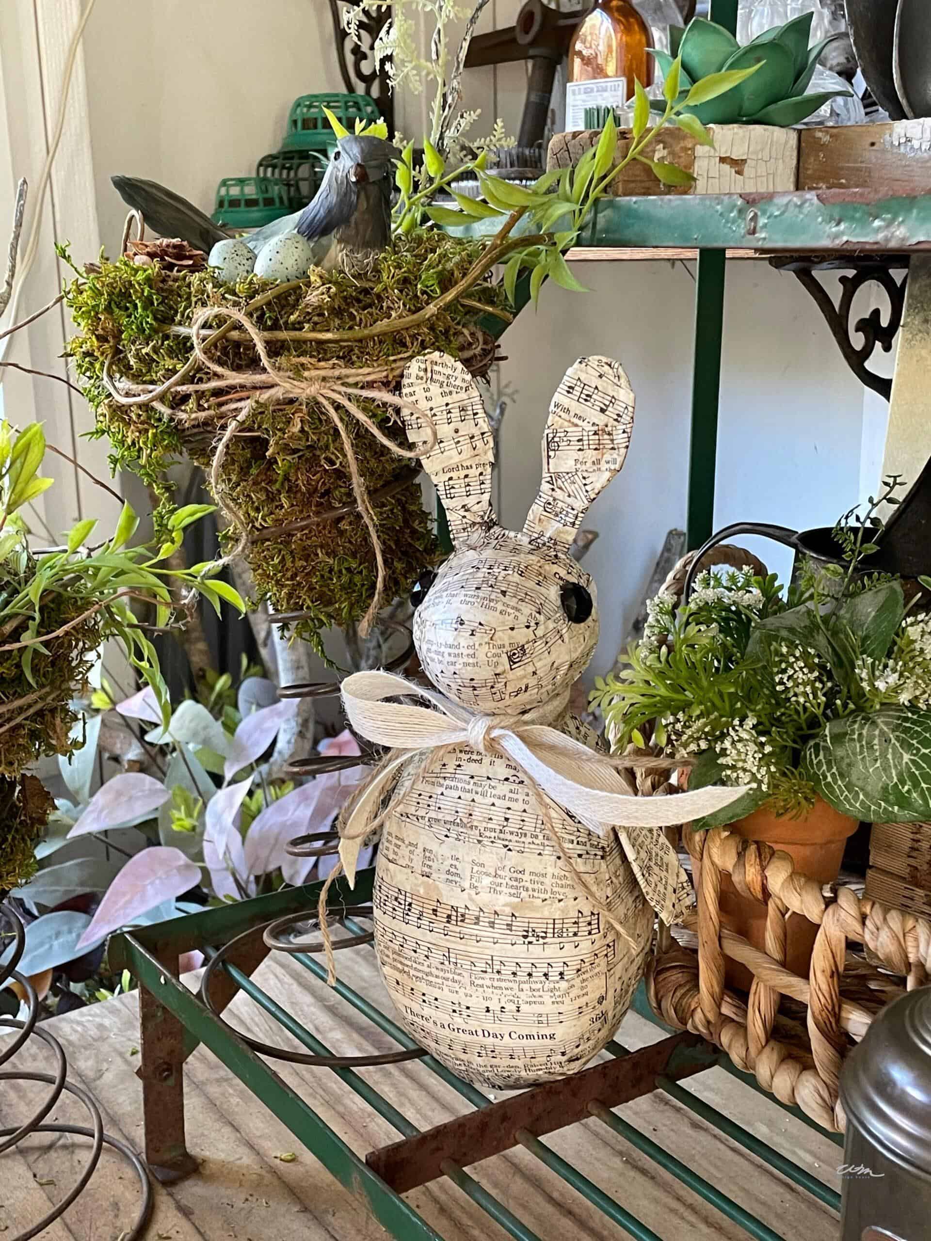 A decorative bunny figurine wrapped in sheet music paper with a ribbon around its neck sits on a shelf surrounded by potted plants, greenery, and garden decor.
