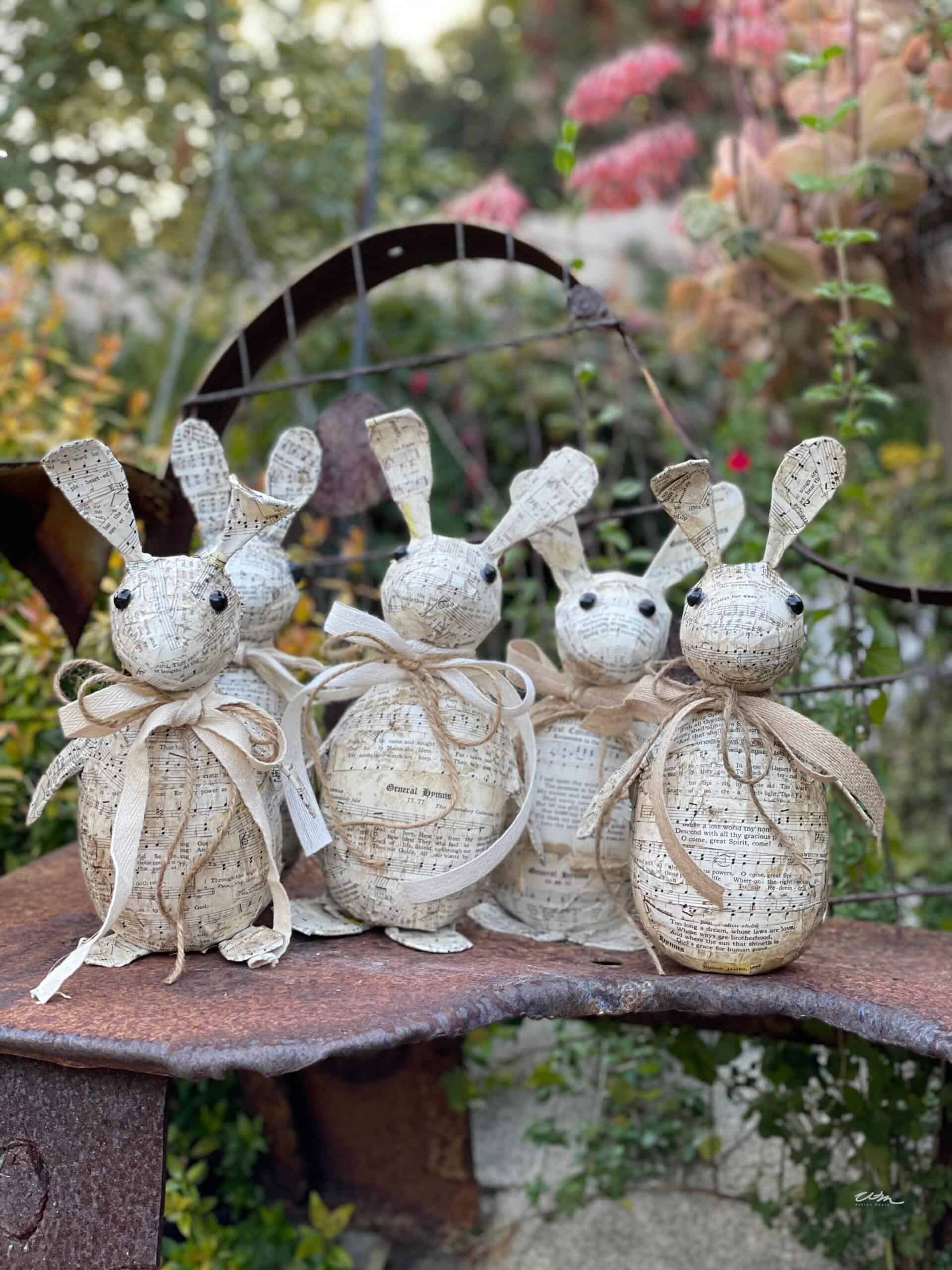 Four rustic bunny figurines with large ears and bows around their necks sit on a weathered metal surface in a garden, surrounded by greenery and pink flowers in the background.