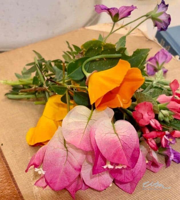 A small bouquet of colorful flowers, including vibrant pink bougainvillea, bright yellow petals, and purple blooms, arranged on a brown surface. The background is slightly blurred and includes a hint of a blue book.