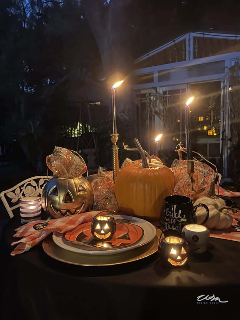 A Halloween-themed table setting at night with lit candles, jack-o'-lanterns, and autumn decorations. Behind the table is a rustic shed adorned with corn stalks and plants. The table features a festive tablecloth, mugs, and plates, creating a cozy, spooky atmosphere.