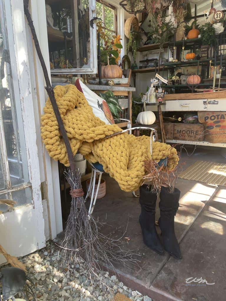 DIY witch's broom made from a large stick and foraged twigs displayed outdoors with some black boots leaned against the door of a gardens shed with pumpkins and other fall decorations