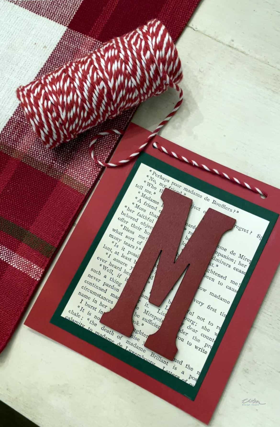 A craft project on a table with a large red letter "M" glued to the center of a page from a book, bordered by a red frame. Next to it is a spool of red and white baker's twine, perfect for creating a Merry Christmas banner. The table has a red plaid tablecloth.