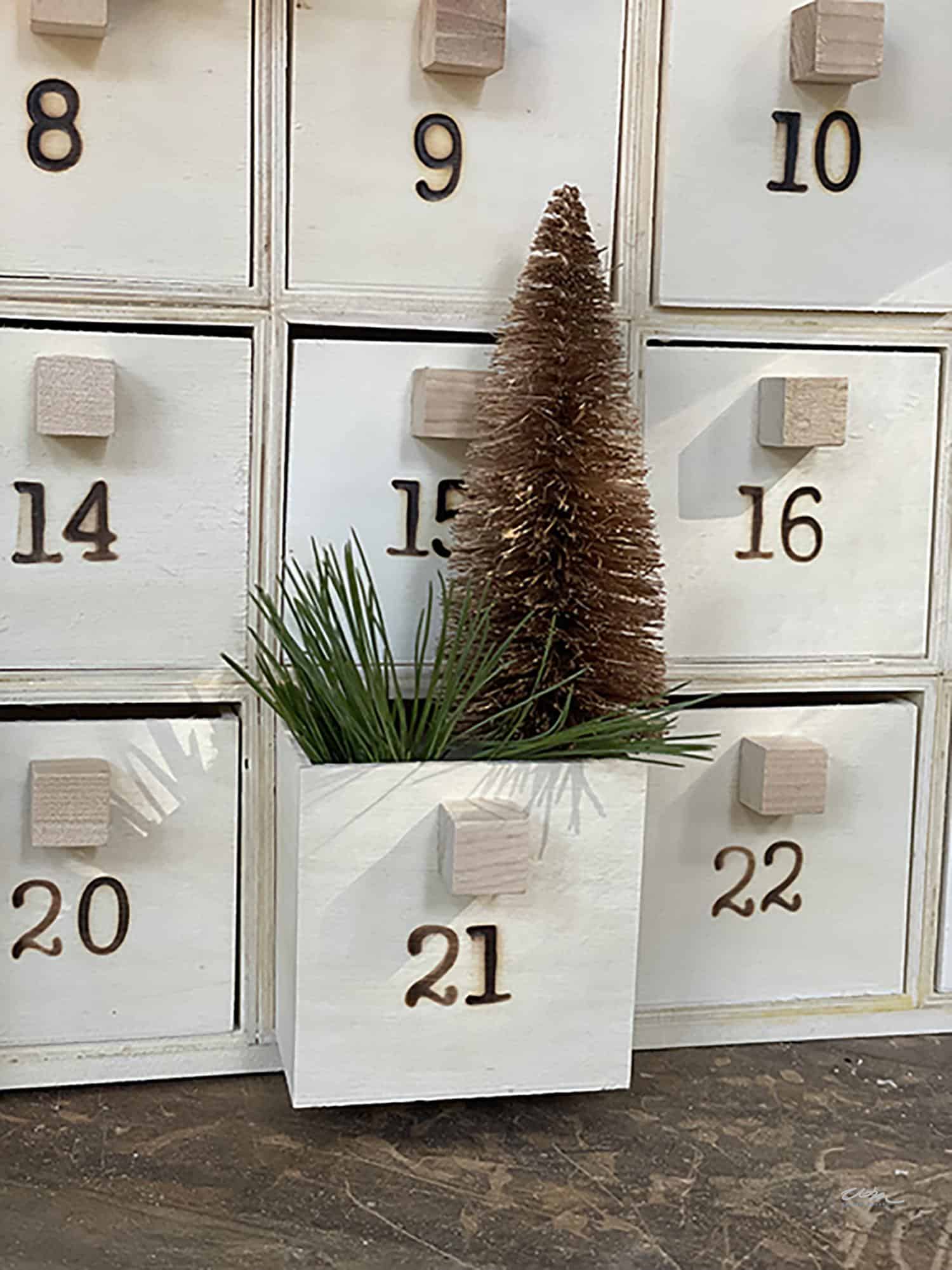 A close-up of a Countdown Advent Calendar for Christmas made with wood boxes with numbered drawers. Drawer 21 is pulled out, revealing pine sprigs and a brown bottle brush tree inside. The drawers around it are numbered 9, 10, 14, 15, 20, and 22.