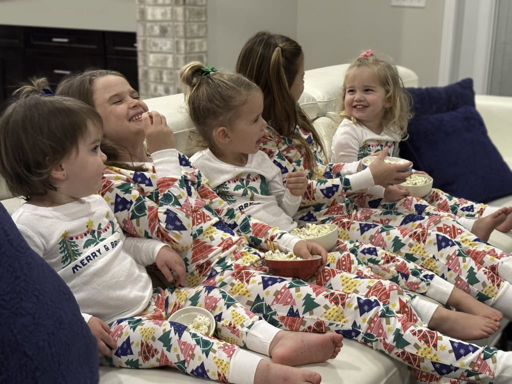 Grandkids sitting on the couch in matching Christmas pajamas laughing and eating popcorn