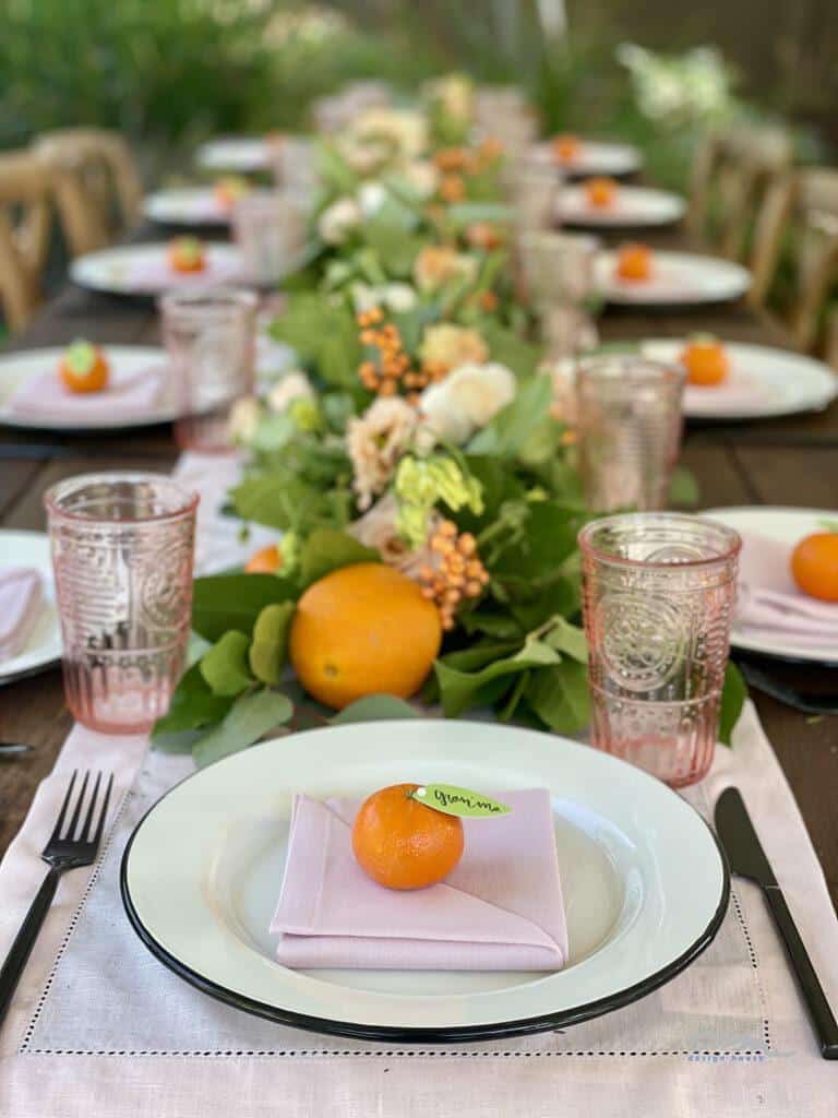 A long, outdoor dining table is set with white plates, pale pink napkins, and oranges on each plate for a Little Cutie Baby Shower. The table is decorated with a lush green garland with white flowers and additional oranges. Pink patterned glasses and silverware complete the charming setting.