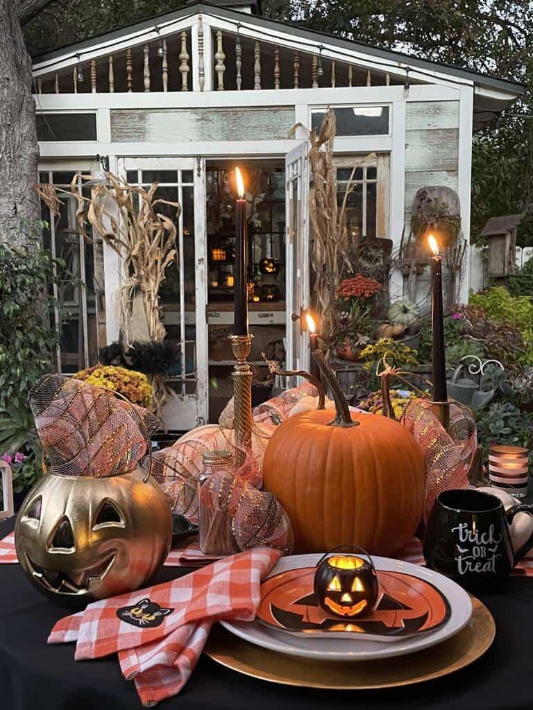 A Halloween-themed table setting with lit candles, jack-o'-lanterns, and autumn decorations. Behind the table is a rustic shed adorned with corn stalks and plants. The table features a festive tablecloth, mugs, and plates, creating a cozy, spooky atmosphere.