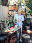 A woman smiles while planting a small plant in a white pot in her garden. She stands at a table with gardening tools, terracotta pots, and flowers. Behind her, there are shelves with more gardening supplies. The setting is bright and filled with greenery.