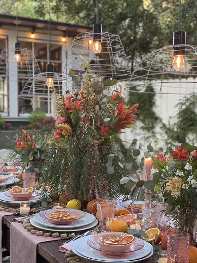 Outdoor dining table with plates, glasses, candles, sliced citrus, and large floral centerpieces; handmade basket pendant light DIY fixtures glow overhead, creating a warm, inviting atmosphere.