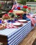 A festive outdoor table set for a patriotic celebration, showcasing Memorial Day table decorations. The setup features a blue and white checkered tablecloth, red and white flowers, an American flag, and an assortment of food, including sandwiches and fruit. Blue plates and woven placemats complete the display.
