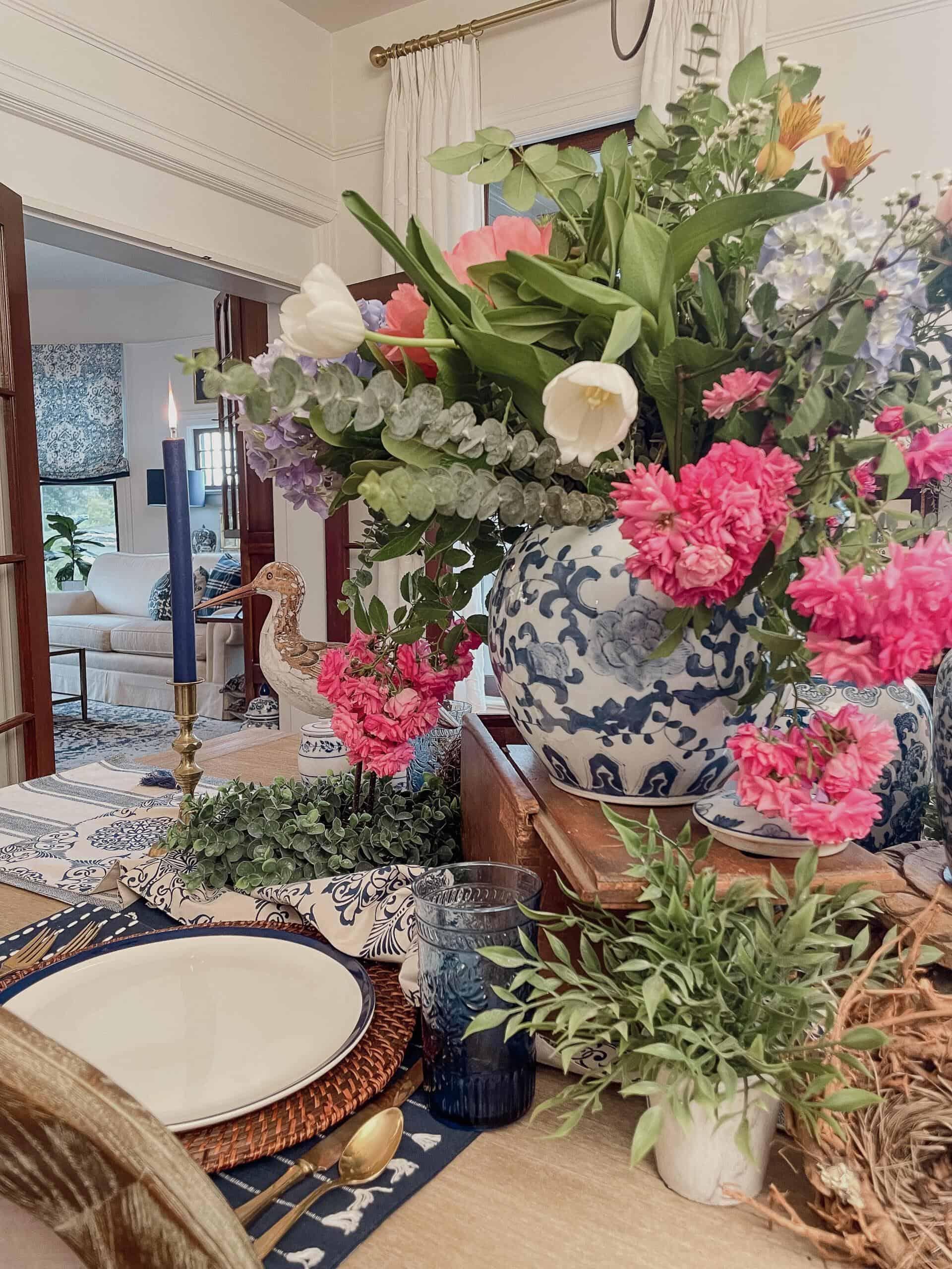 A dining table decorated with a large blue and white vase filled with colorful flowers, surrounded by greenery, a candlestick, a place setting, and blue glassware in a cozy, elegant room.