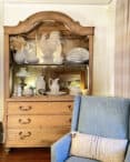 A wooden hutch, its surfaces meticulously stripped with oven cleaner, displays various white ceramic dishes and decor items, including a rooster figurine and a lamp. Beside the hutch is a blue upholstered chair adorned with a pillow that reads "HOME." The room has light-colored walls and partially visible curtains.