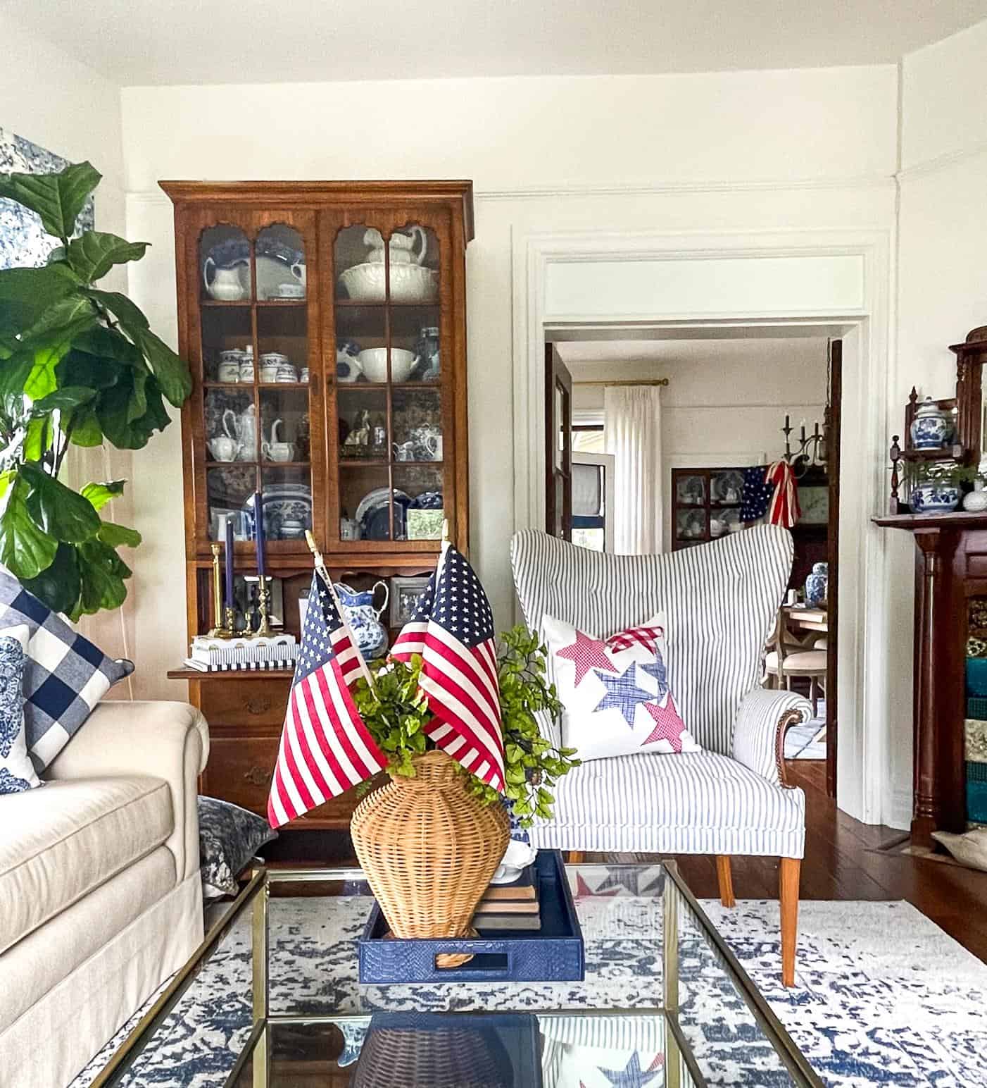 A cozy living room with a glass coffee table holding a wicker basket with two small American flags, showcasing DIY Fourth of July decorations. In the background, there is a wooden cabinet filled with dishes, a striped armchair with star-patterned pillows, and a couch with plaid pillows.