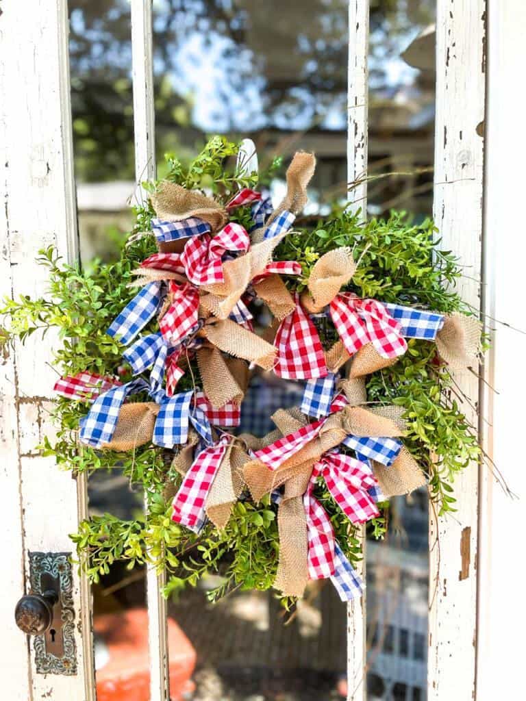 A festive wreath hangs on a white, rustic door. The wreath is adorned with green foliage and decorated with bows made of red, white, and blue checkered and burlap ribbons, creating a cheerful and patriotic appearance.