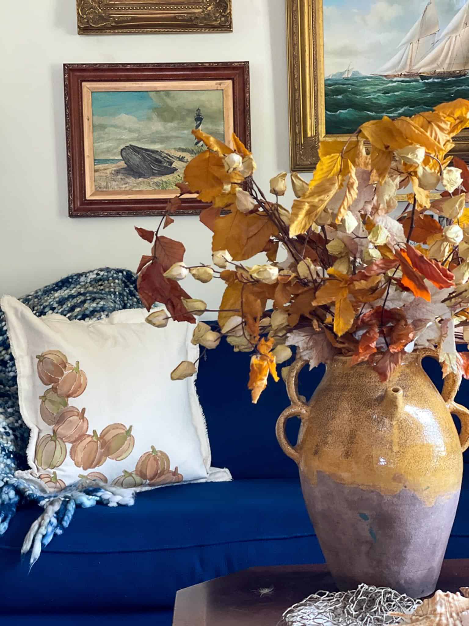 A cozy living room features a dark blue couch adorned with fall throw pillows, including a white one decorated with floral art. Above the couch, various framed paintings hang on the wall. In the foreground, a large vase holds an arrangement of autumnal leaves and dried branches.