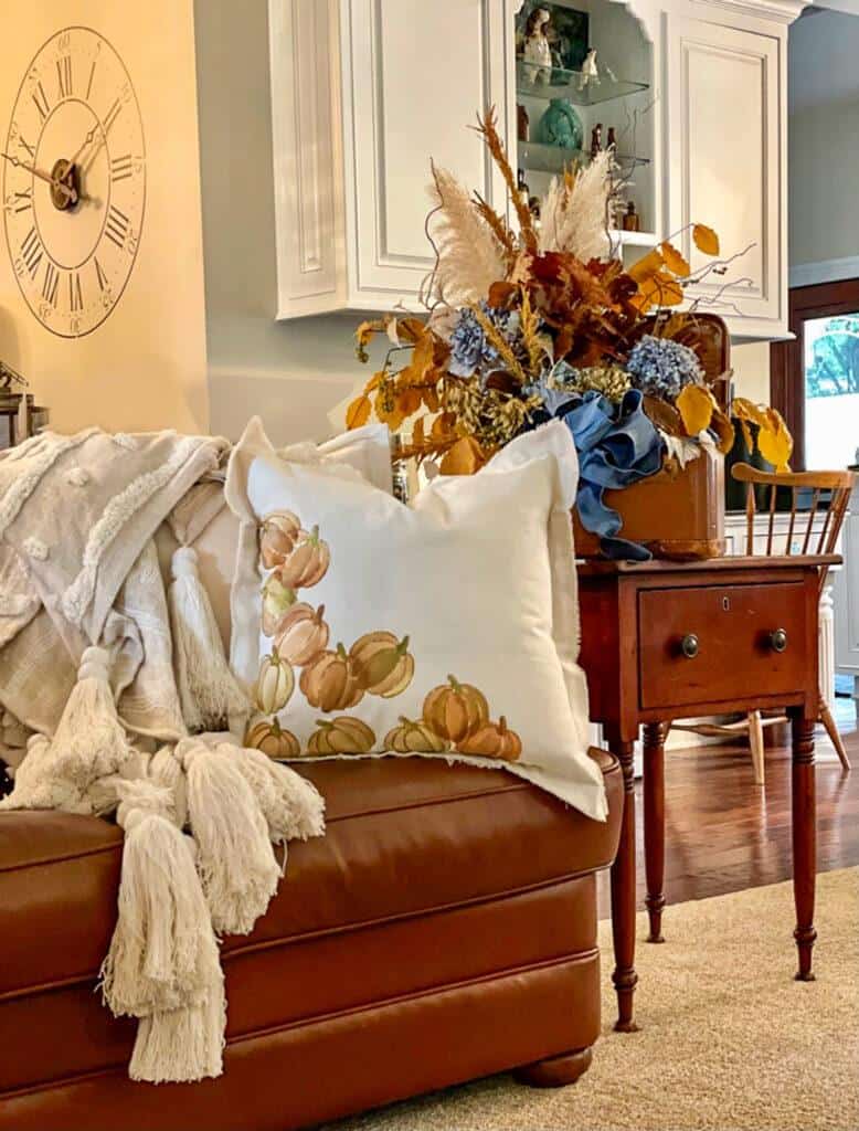 A cozy living room scene featuring a brown leather couch adorned with a white throw blanket and fall throw pillows displaying pumpkin designs. A wooden side table holds an autumn-themed floral arrangement with blue, white, and orange elements. A large wall clock is visible in the background.