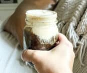 A hand holds a mason jar filled with a dark beverage topped with a frothy cream layer. The background shows a light-colored knitted blanket and a brown cushion on a sofa. The setting suggests a cozy indoor environment.