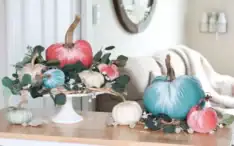 A centerpiece on a table features a mix of pink, blue, white, and green pumpkins, adorned with green leaves and white flowers. Behind it, a cozy living room with a beige sofa and a blanket is partially visible. A mirror and wall decor add a homely touch to the backgroundโperfect for our Fabulous Friday Link Party on 8.19.22!