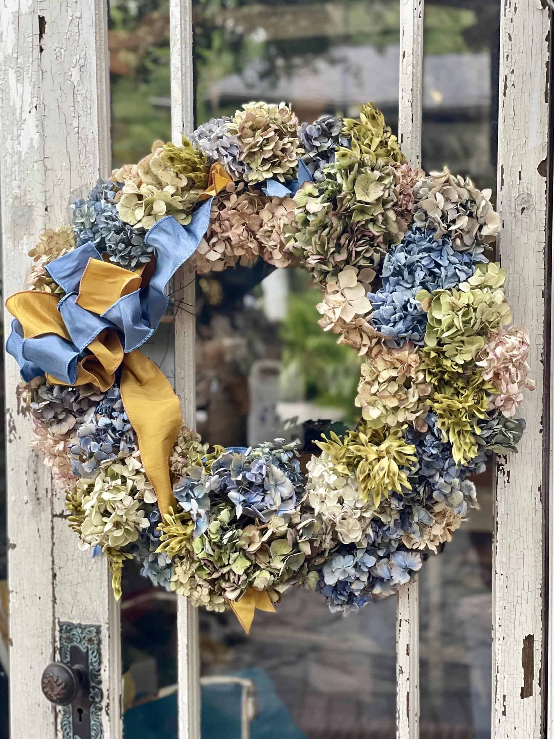 A wreath made of dried hydrangeas in pastel shades of blue, green, and beige hangs on a rustic, distressed white door. The wreath is adorned with a blue and yellow ribbon.