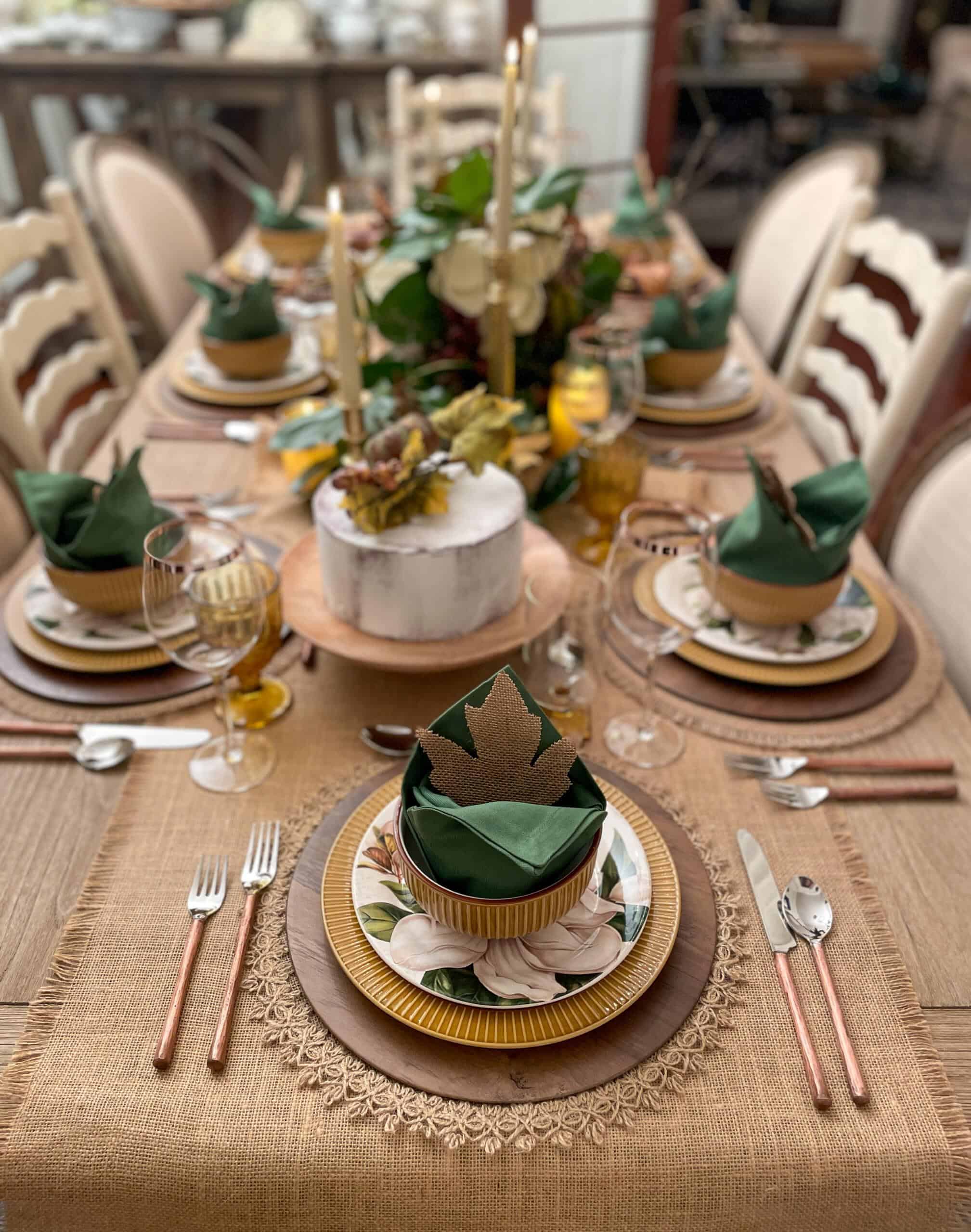 A beautifully set rectangular dining table, graced with a rustic beige tablecloth, adorned with green and gold napkins shaped like leaves. Various plates, gold utensils, wine glasses, and a centerpiece of candles and a white cake surrounded by floral decorations complete this perfect Amazon Fall Decor setting.