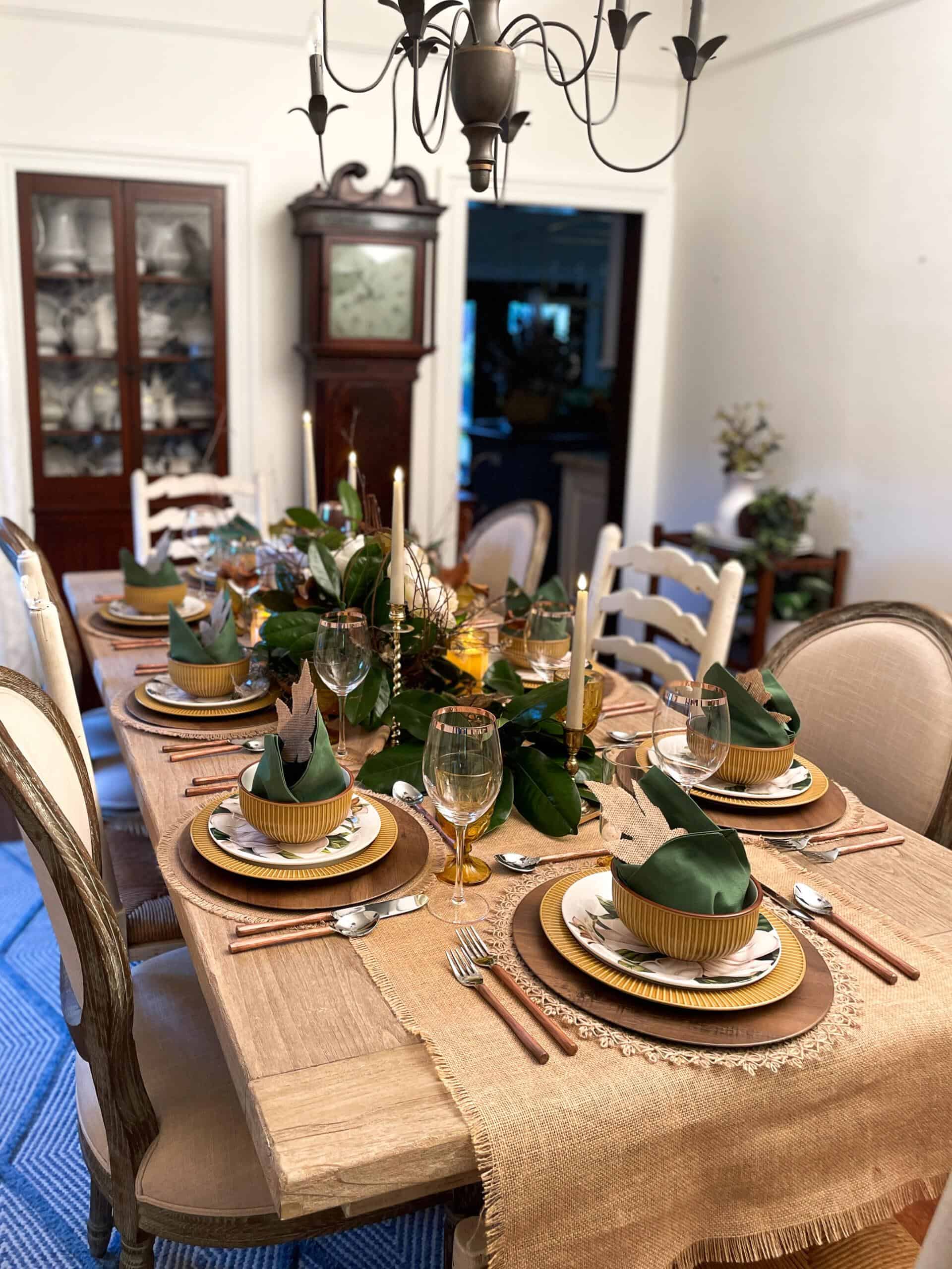 A dining table elegantly set for a meal, adorned with green and gold tableware, green napkins, and gold-rimmed glasses. A beige table runner with fringed edges, a centerpiece with foliage and candles, and a chandelier overhead complete the sophisticated Amazon Fall Decor-inspired setting.