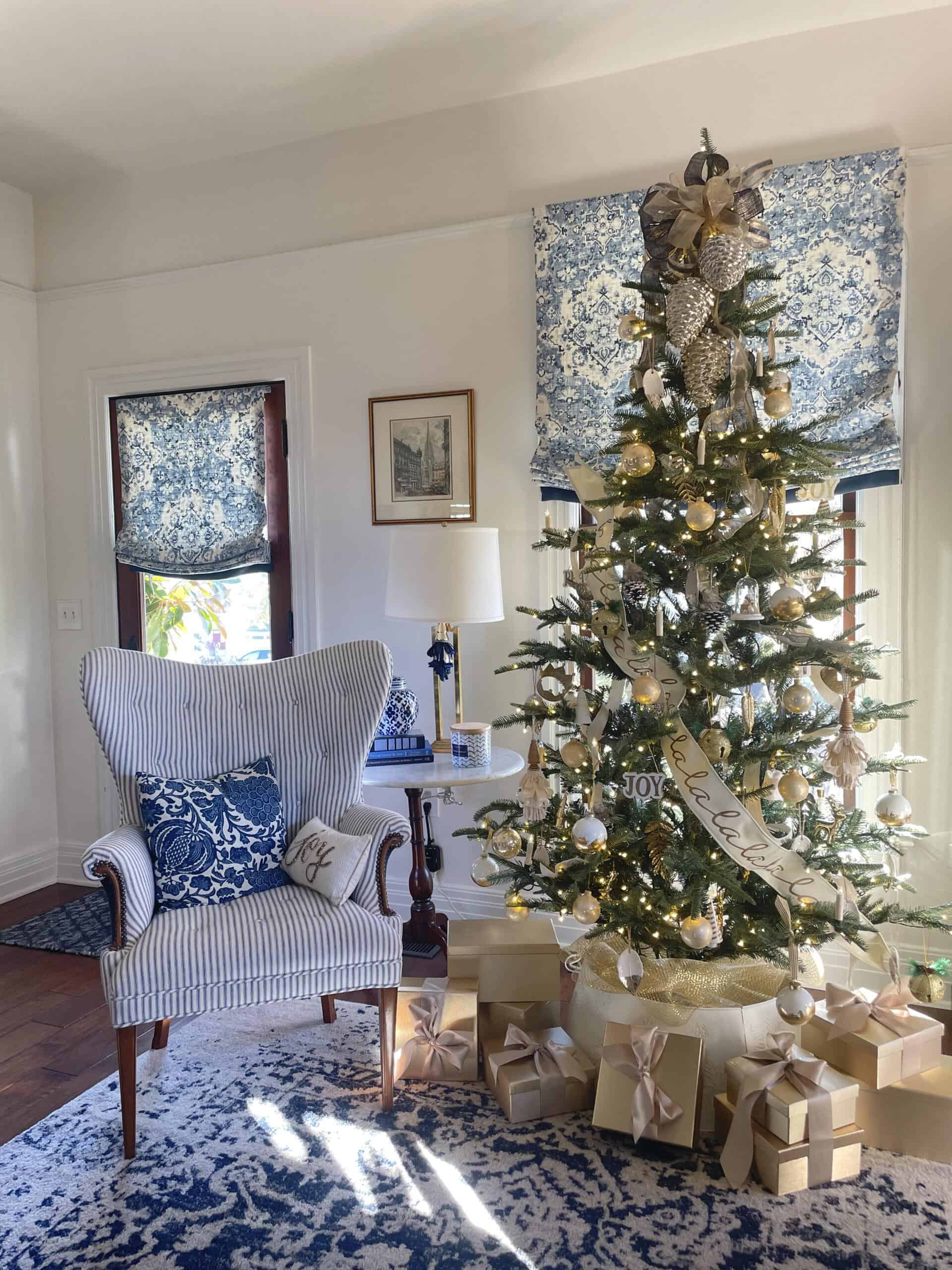 A decorated Christmas tree, the King of Christmas Trees, with gold, silver, and white ornaments. Wrapped presents sit underneath. To the left, a striped armchair with blue and white pillows is near a small wooden table and lamp in a room adorned with blue floral accents.