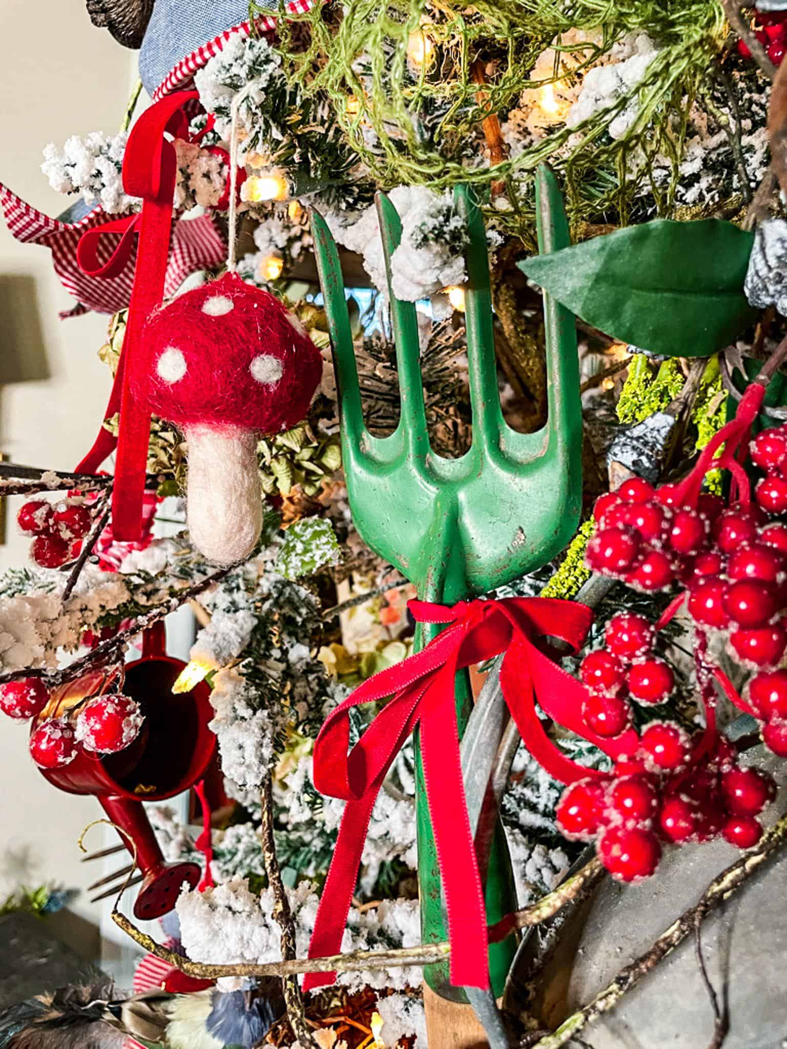 A Christmas tree decorated with a green garden fork adorned with a red ribbon, a felt red mushroom with white spots, red berry clusters, and other rustic ornaments. The tree is dusted with artificial snow and illuminated by warm white lights.