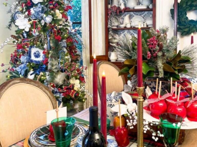 A festive dining room decorated for Christmas. A table is set with red candles, glassware, and a wine bottle. Nearby, a blue and white Christmas tree adorned with ornaments and lights adds charm. A cabinet with white dishes stands against the wall.