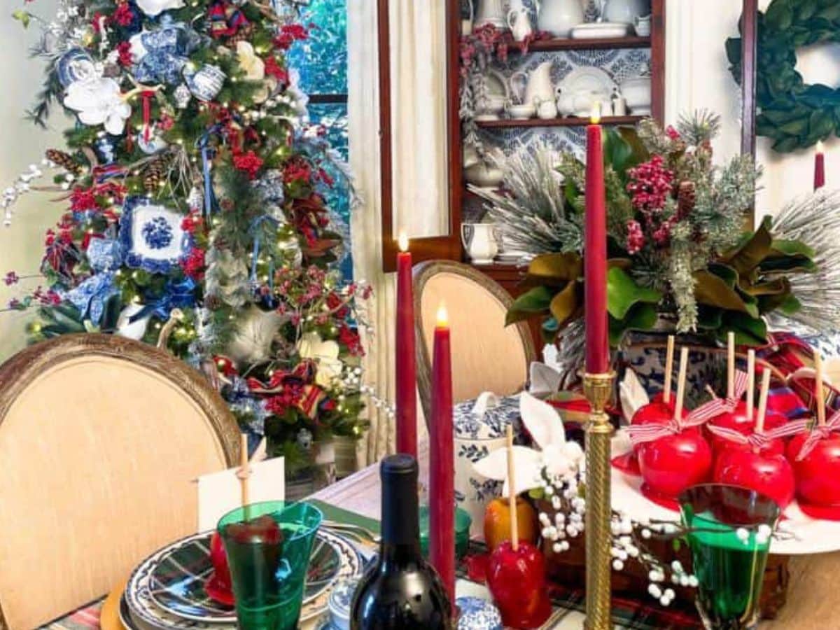 A festive dining room decorated for Christmas. A table is set with red candles, glassware, and a wine bottle. Nearby, a blue and white Christmas tree adorned with ornaments and lights adds charm. A cabinet with white dishes stands against the wall.