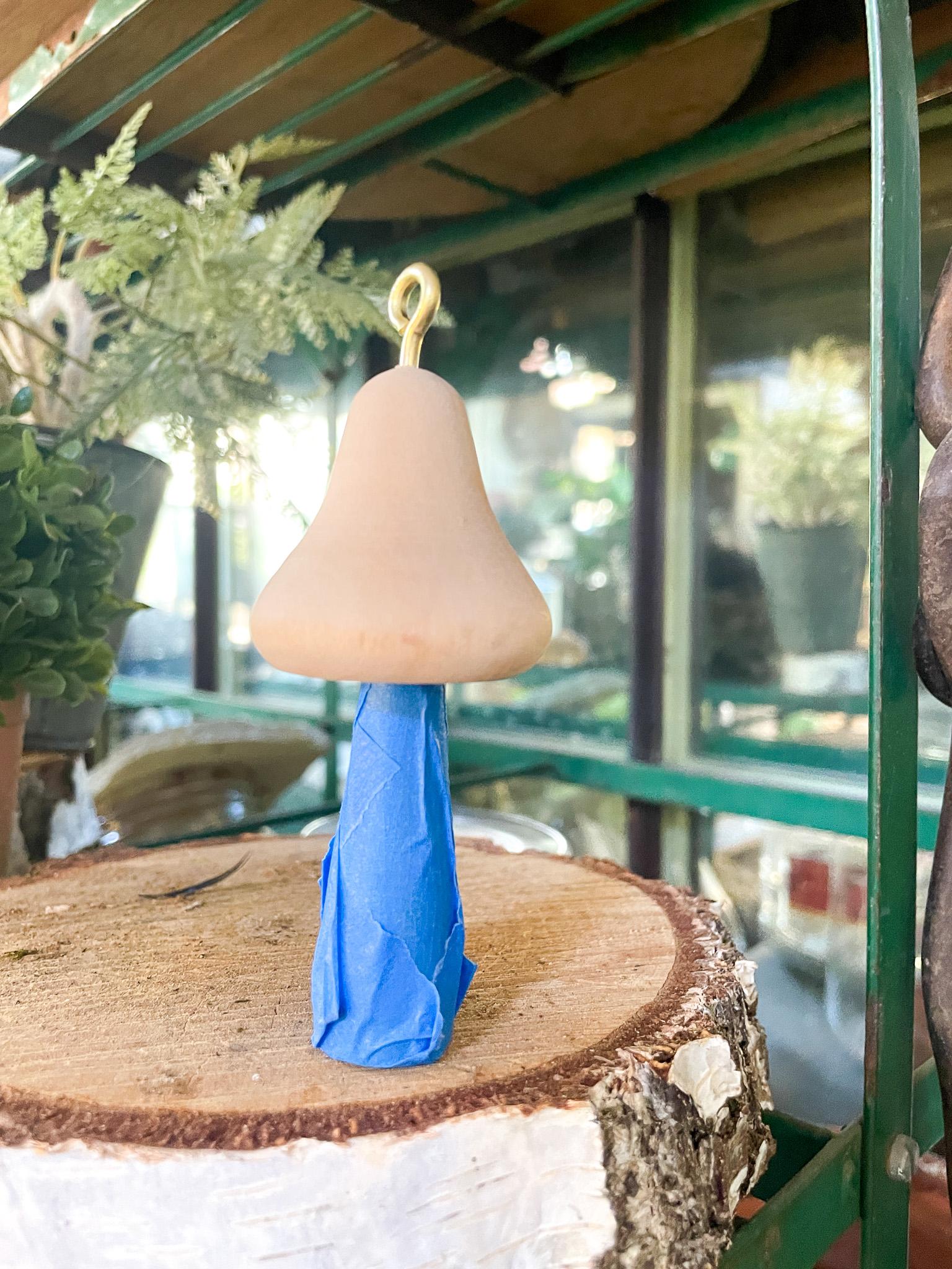 A small clay mushroom with a smooth, beige cap and a blue painted stem is displayed on a tree stump slice. In the background, there are green plants and a glass greenhouse structure. The mushroom has a loop at the top, suggesting it might be a decoration or ornament.