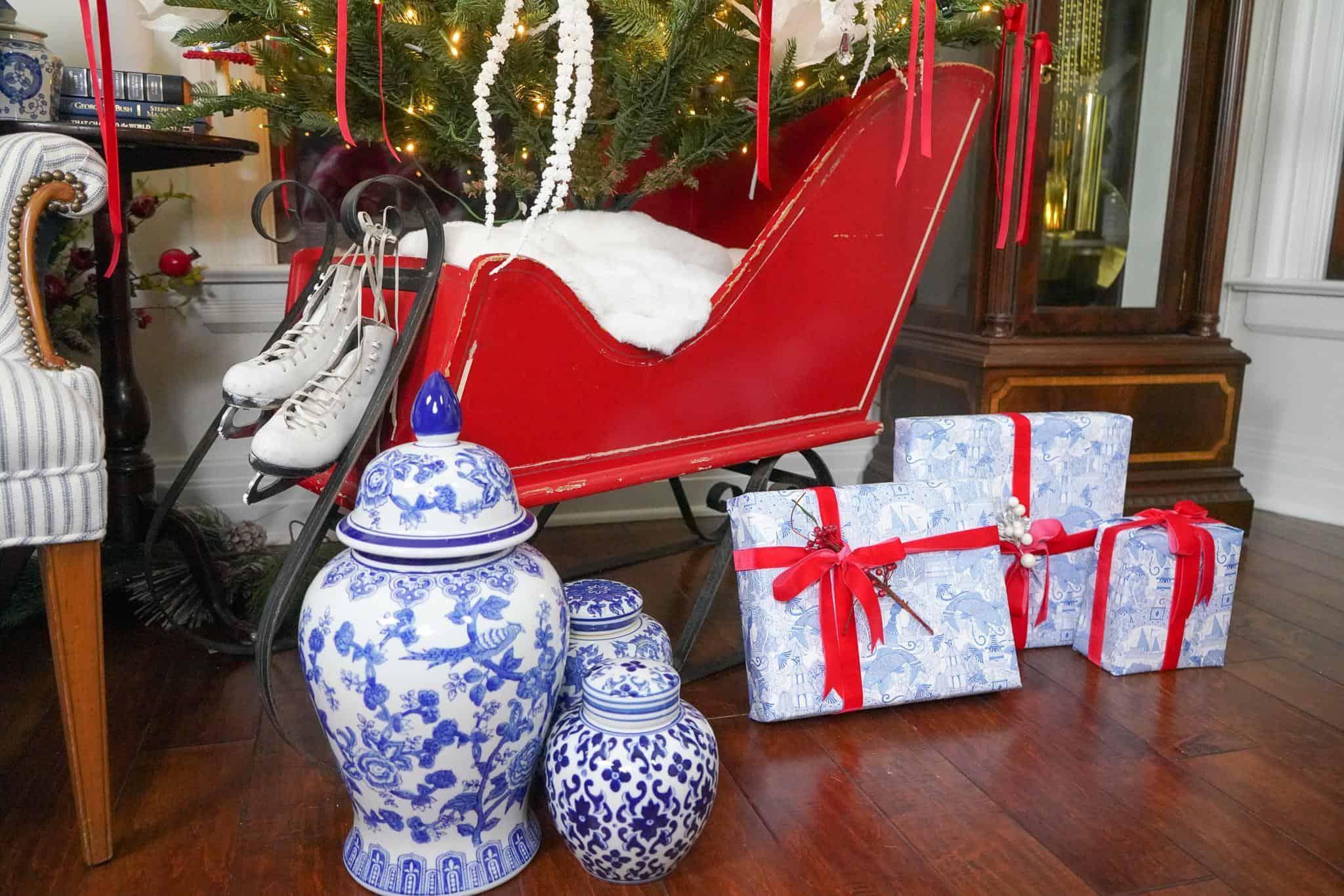 A festive Christmas scene featuring a red sleigh filled with white fabric and adorned with ice skates. Blue and white porcelain vases are on the floor next to wrapped gifts with blue patterns and red ribbons. A decorated Christmas tree is in the background.