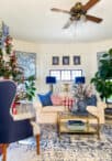 A cozy and elegantly decorated living room features a white sofa adorned with blue and red pillows. A coffee table holds candles and a vase, with books on the lower shelf. Classic Christmas decorations enhance the large Christmas tree in the corner, and various plants add greenery to the room.