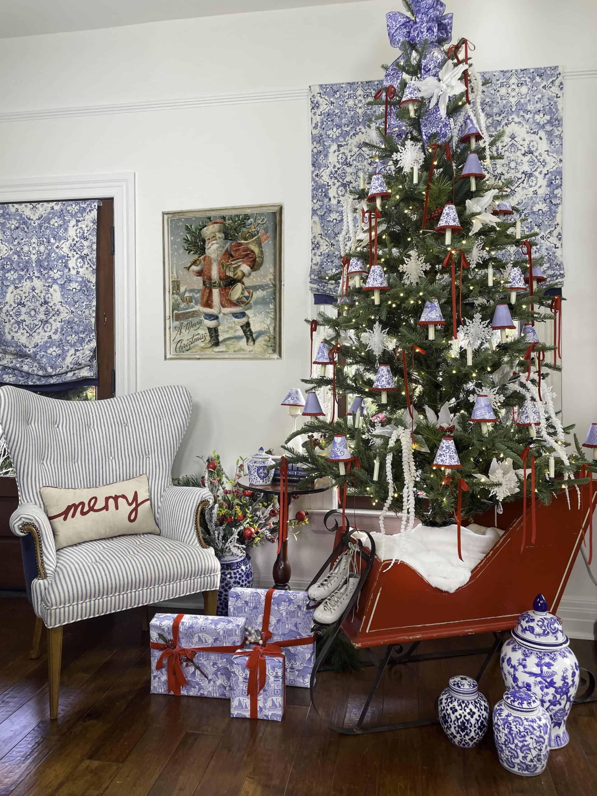 A Christmas tree adorned with purple bells, red ribbons, and white ornaments stands in a red sleigh filled with faux snow. Surrounding the tree are wrapped gifts, a blue and white patterned chair with a "merry" pillow, vases, and festive decorations.