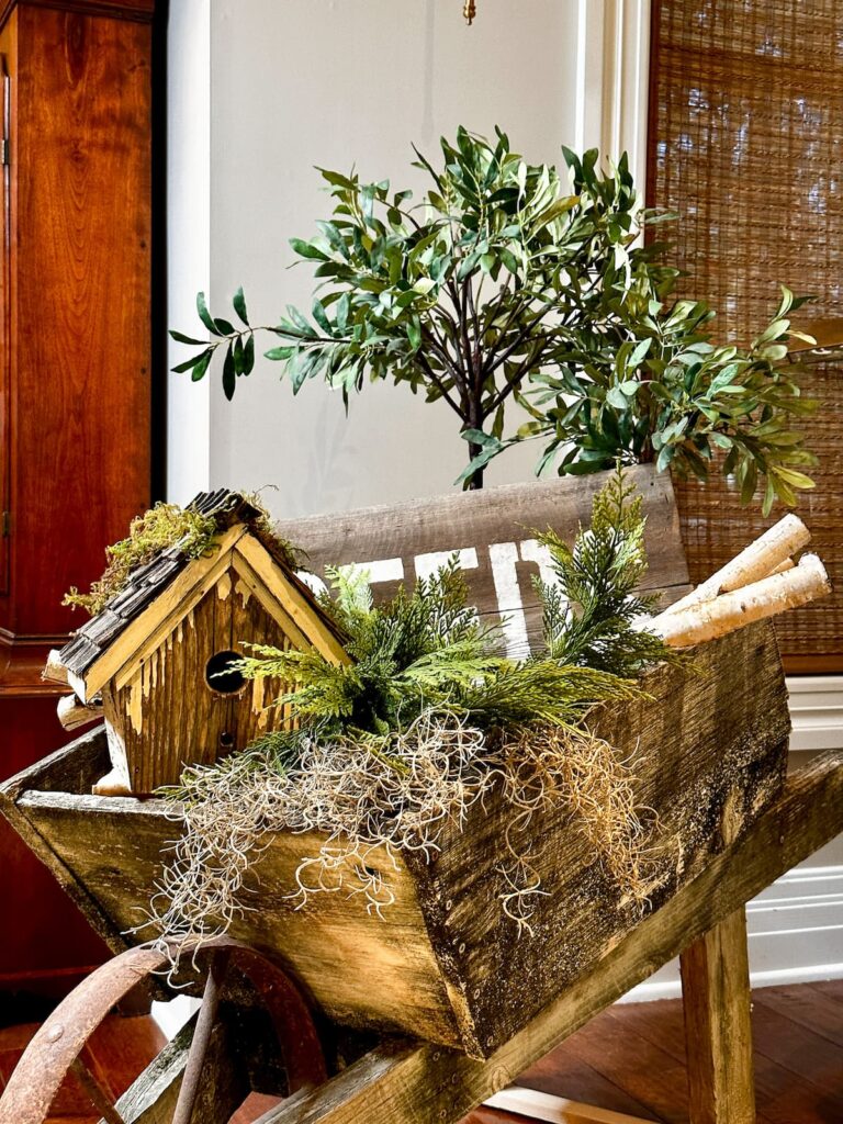 wooden wheel barrel with greenery, moss, birdhouse and olive tree. -Winter floral arrangement