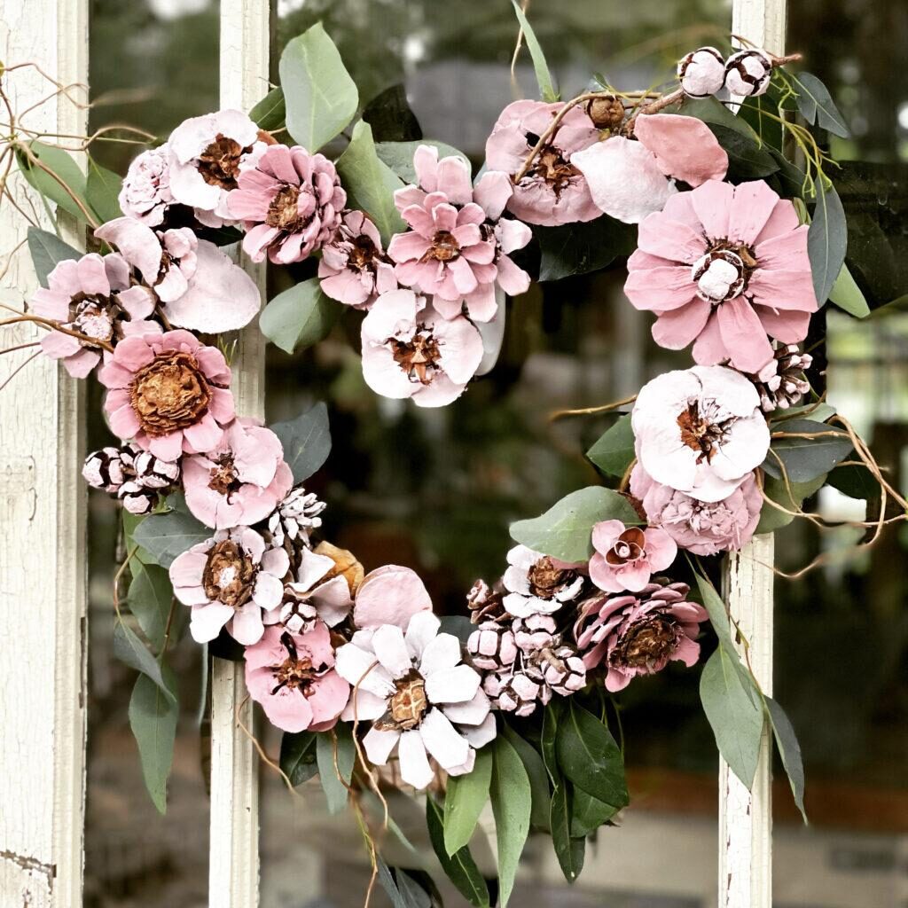 A heart-shaped wreath made of pinecones and white, pink, and brown flowers, interspersed with green leaves, is hanging on a wooden door. The background shows a blurred view of greenery.