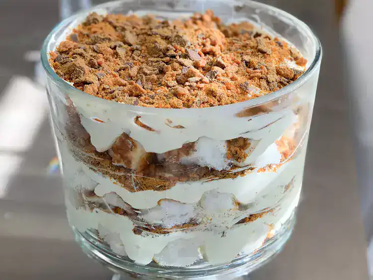 A glass dish filled with layered dessert, featuring creamy filling and a crumbled brown topping—likely made with store-bought angel food cake—on a light-colored surface.