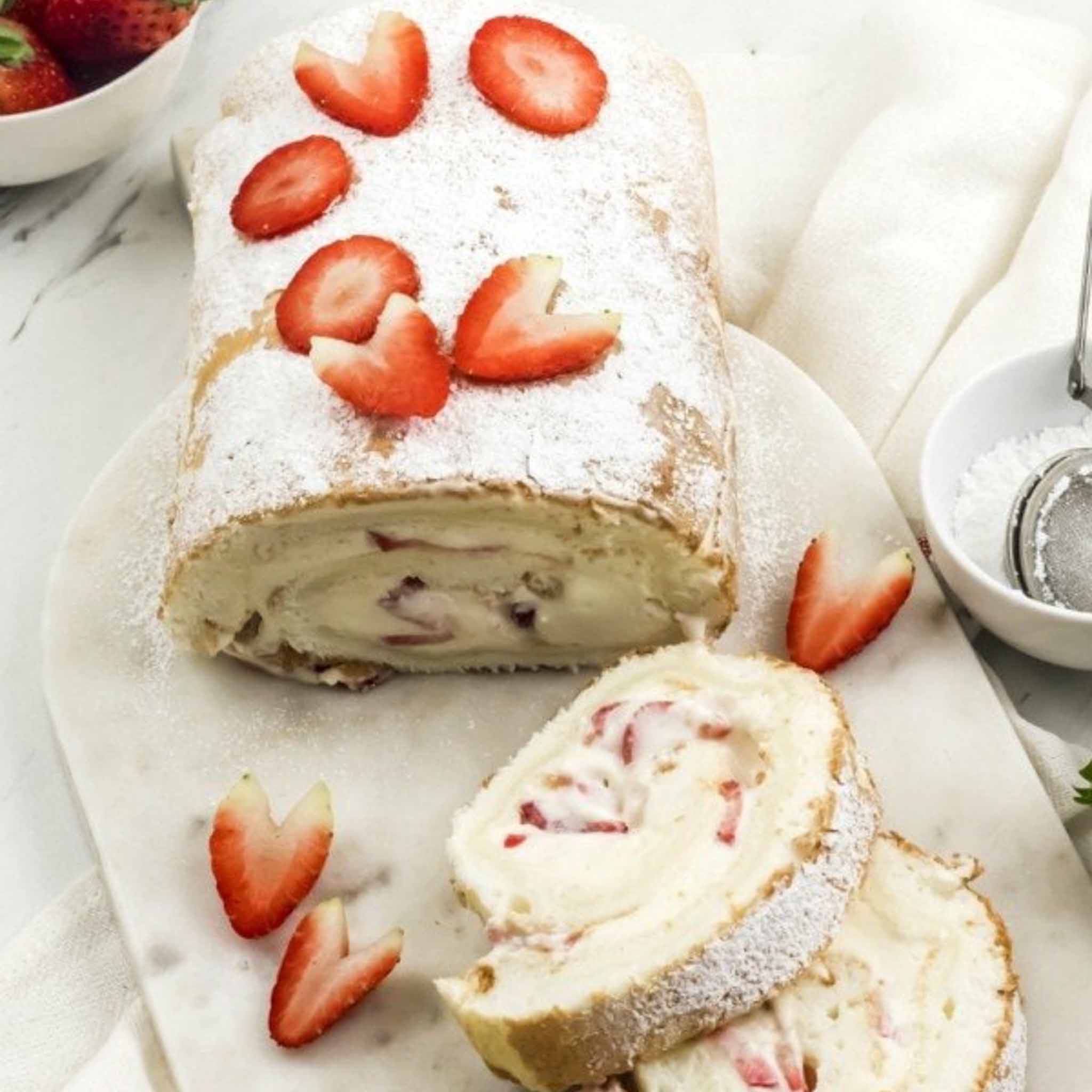 A sliced strawberry Swiss roll cake, made with store-bought angel food cake and topped with powdered sugar and fresh strawberry pieces, is displayed on a marble board, with a bowl of strawberries and a sifter nearby.