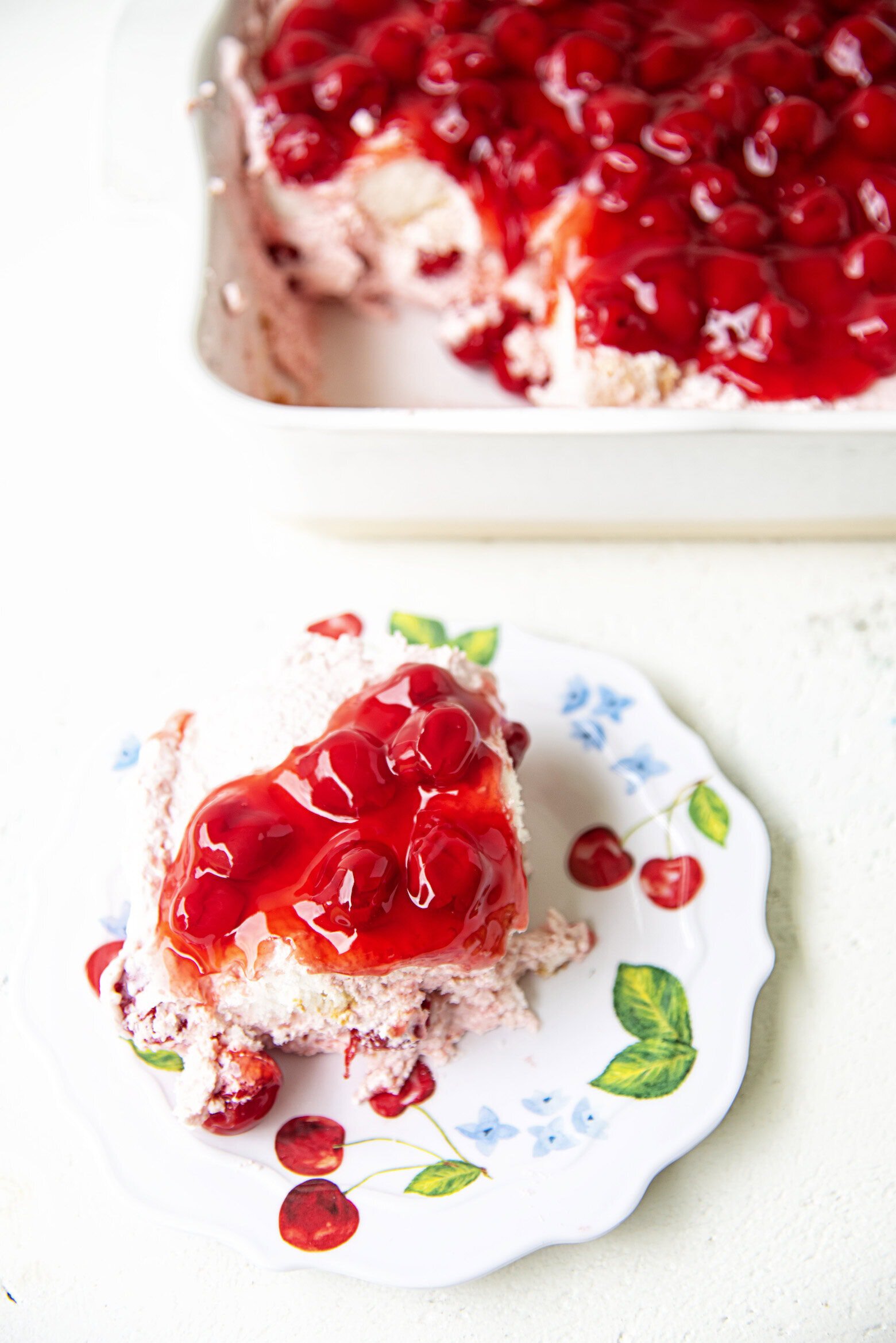 A slice of cherry dessert made with store-bought angel food cake and crowned with whipped topping and cherries sits on a floral plate, in front of a baking dish filled with more of the cherry-topped treat.