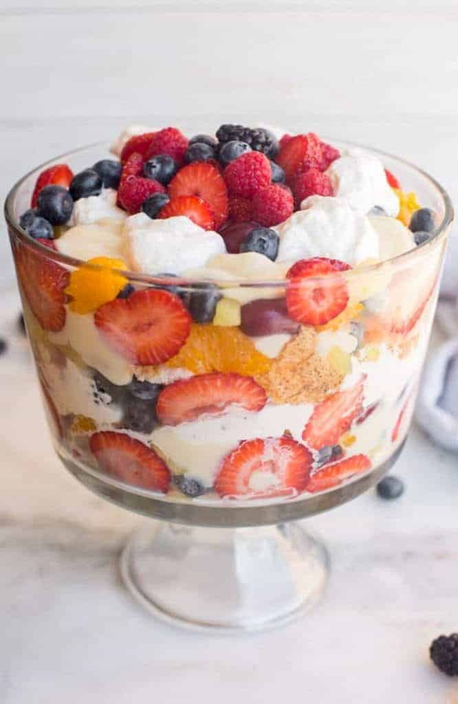 A glass trifle bowl filled with layers of strawberries, blueberries, blackberries, whipped cream, cubes of store-bought angel food cake, and custard, topped with mixed berries and dollops of whipped cream.