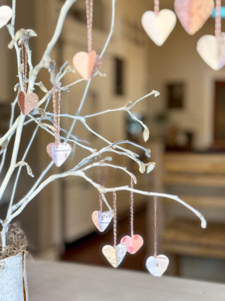 A white, bare branch displayed in a pot is decorated with various paper hearts hanging from red-and-white striped strings. The hearts are in different colors and patterns, adding a whimsical touch to the scene. The background is softly blurred.