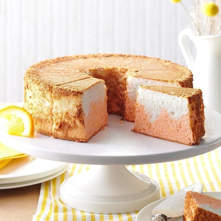 A two-tone orange and white chiffon cake, reminiscent of a store-bought angel food cake, sits on a white cake stand with one slice cut out. A plate with an orange slice garnish is nearby on a yellow and white striped tablecloth.