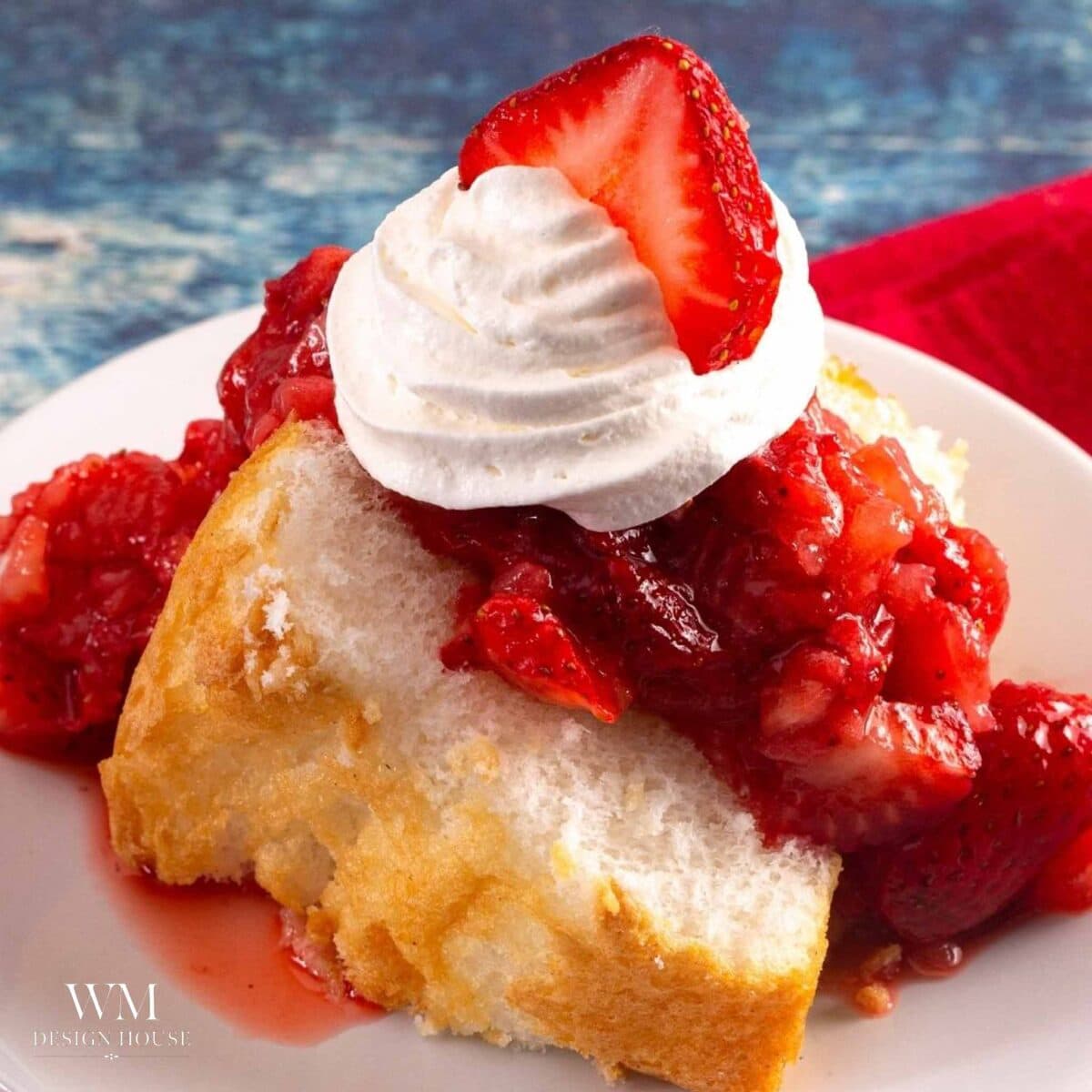 A slice of store-bought angel food cake topped with chopped strawberries in syrup, a swirl of whipped cream, and a fresh strawberry slice, served on a white plate.