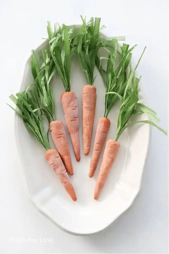 Six realistic carrot-shaped objects with green paper tops are neatly arranged on a white oval dish. The carrots appear to be made out of a material resembling wood or clay, perfect for decorating your She-Shed. The white background creates a clean and minimalistic presentation.