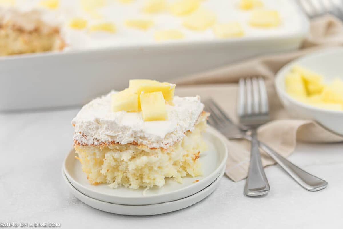 A slice of white cake made with store-bought angel food cake, topped with whipped topping and pineapple chunks, sits on a plate; a baking dish and more cake are in the background.
