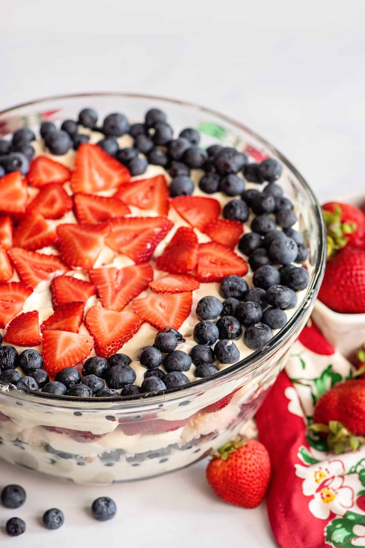 A glass punch bowl of layered trifle made with store-bought angel food cake, topped with sliced strawberries arranged in a star shape and surrounded by blueberries. A red floral napkin and whole strawberries are beside the bowl.